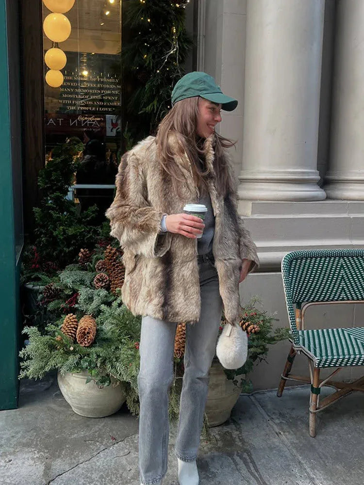 Woman wearing a brown and tan faux fur jacket, grey jeans, and a green baseball cap, holding a coffee cup and a fluffy white purse.