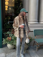 Woman wearing a brown and tan faux fur jacket, grey jeans, and a green baseball cap, holding a coffee cup and a fluffy white purse.