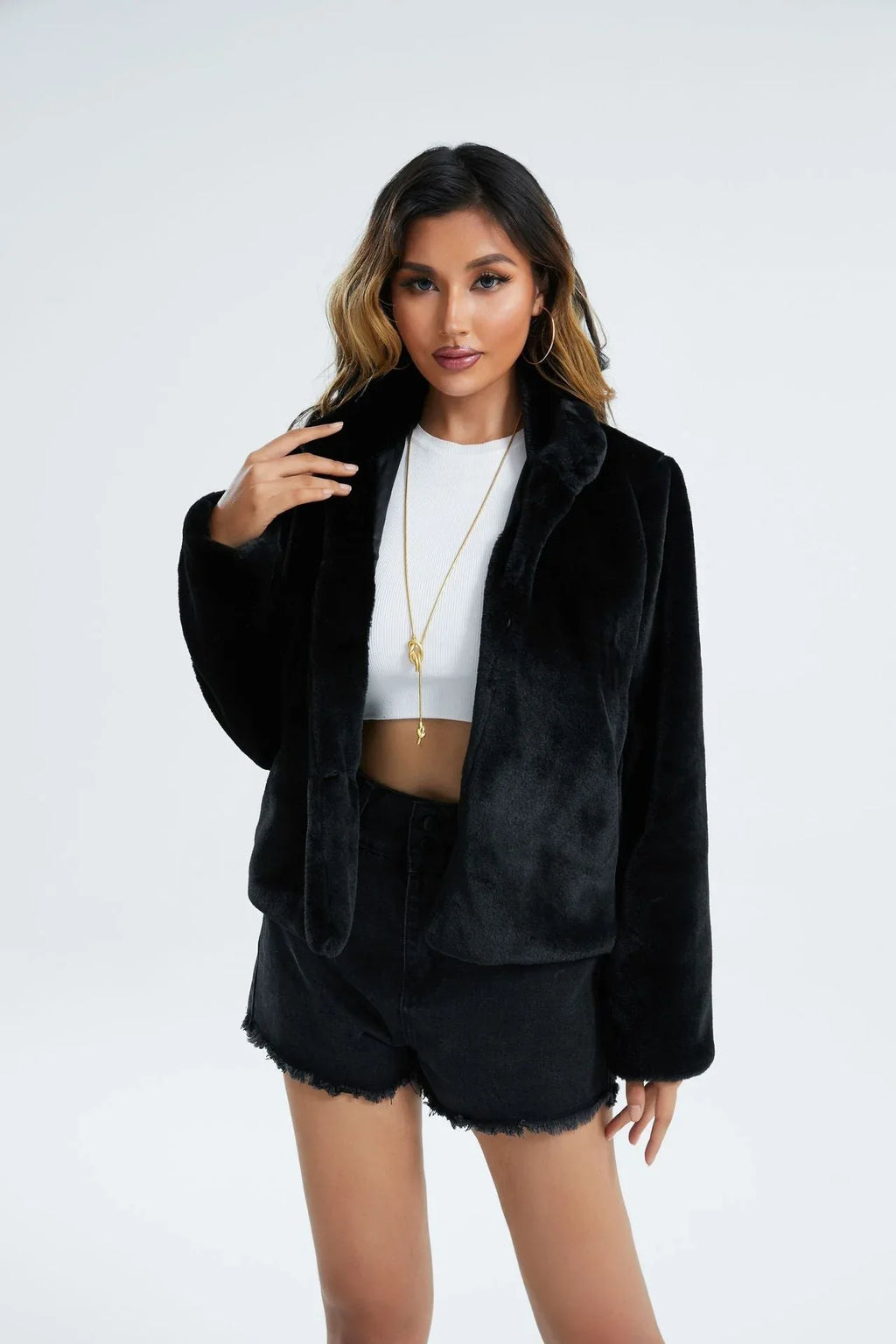 Woman wearing a black faux fur cropped jacket over a white top and black denim shorts, touching her collar.