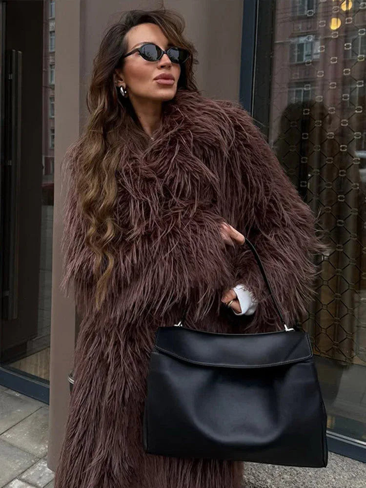 Close-up of a woman wearing a long brown shaggy faux fur coat, sunglasses, and silver earrings, holding a black handbag.