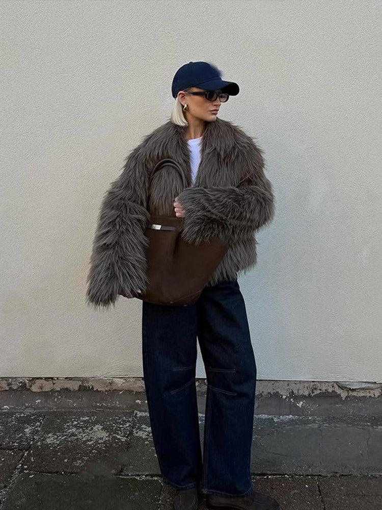 Woman in a brown shaggy faux fur jacket, dark wash wide-leg jeans, and a navy baseball cap, standing against a textured wall.