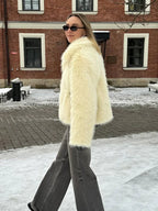 Side view of a woman in a cream faux fur jacket and grey wide-leg jeans walking in a snowy environment.