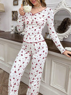 Woman leaning against a dresser, drinking tea, wearing white pajamas with red floral print and scalloped trim.
