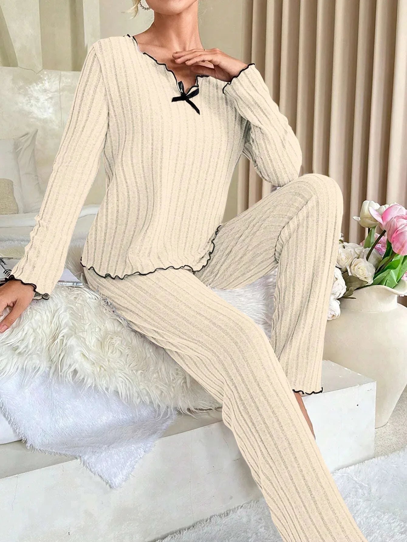 Woman sitting on a bed wearing a cream-colored ribbed knit loungewear set with black trim and a black bow, touching her neck.