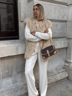 Woman wearing a tan faux fur vest over a white cable-knit sweater and white wide-leg pants.