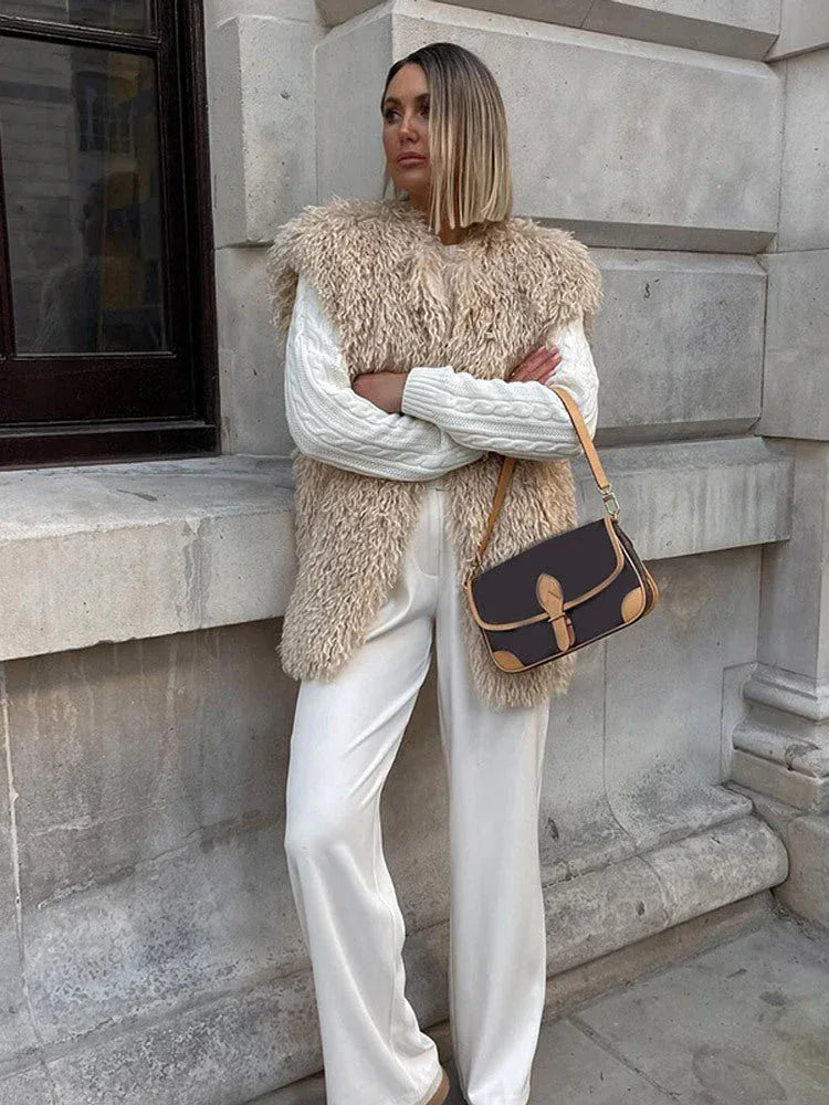 Woman wearing a tan faux fur vest over a white cable-knit sweater and white wide-leg pants.
