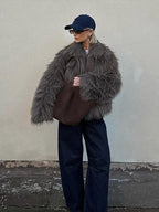 Full body shot of a woman wearing a brown shaggy faux fur jacket, dark wash wide-leg jeans, and a navy baseball cap, standing with her side to the camera.