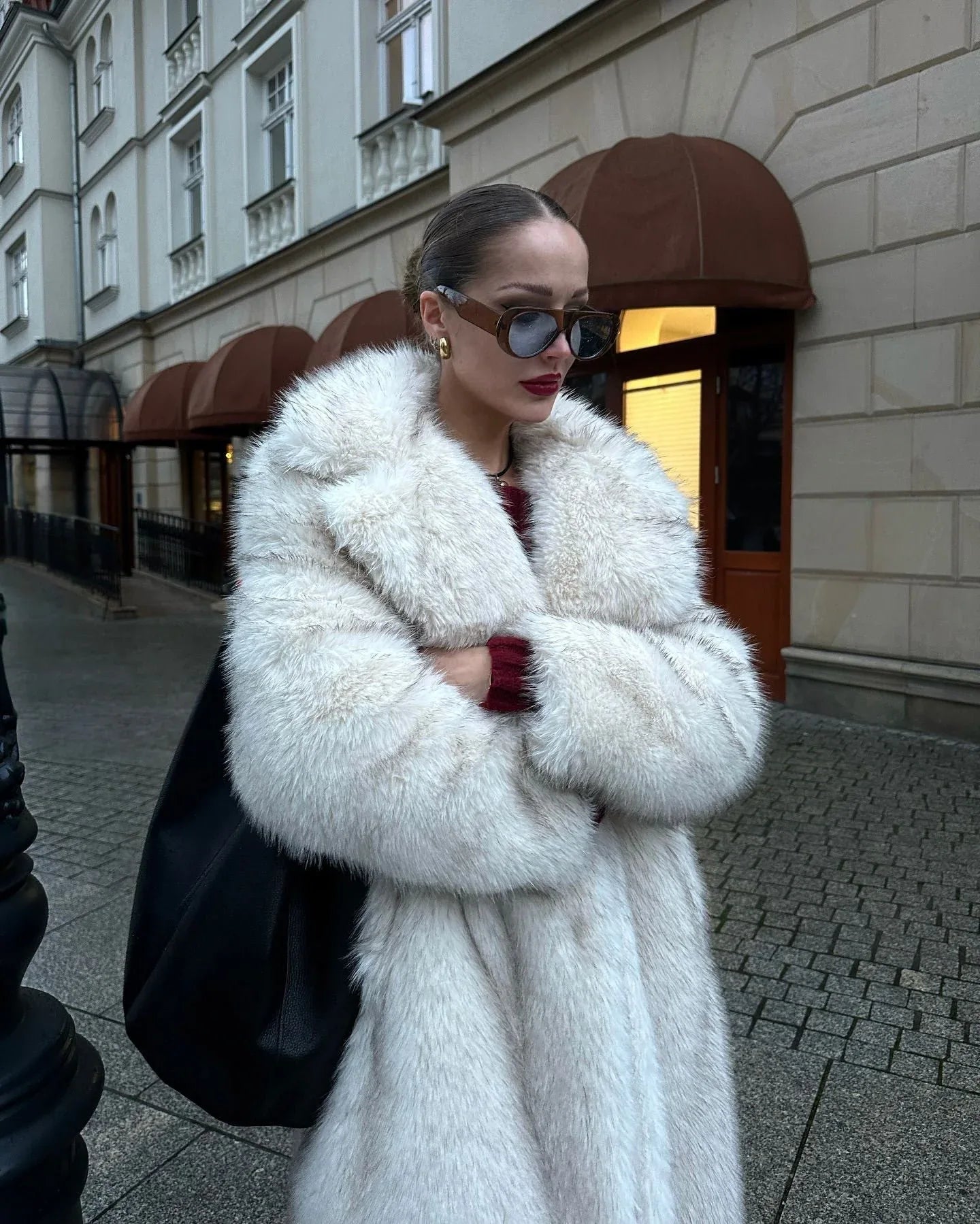 Woman in a white faux fur coat with her arms crossed, wearing sunglasses and gold earrings.