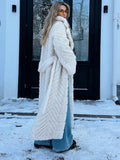 Rear view of a woman wearing a cream faux fur longline coat with chevron texture and a belted waist, standing in front of a building with snow on the ground.