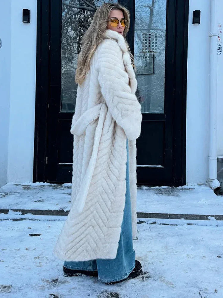 Rear view of a woman wearing a cream faux fur longline coat with chevron texture and a belted waist, standing in front of a building with snow on the ground.