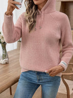 Woman holding a phone, wearing a pink hooded knit sweater and blue denim jeans