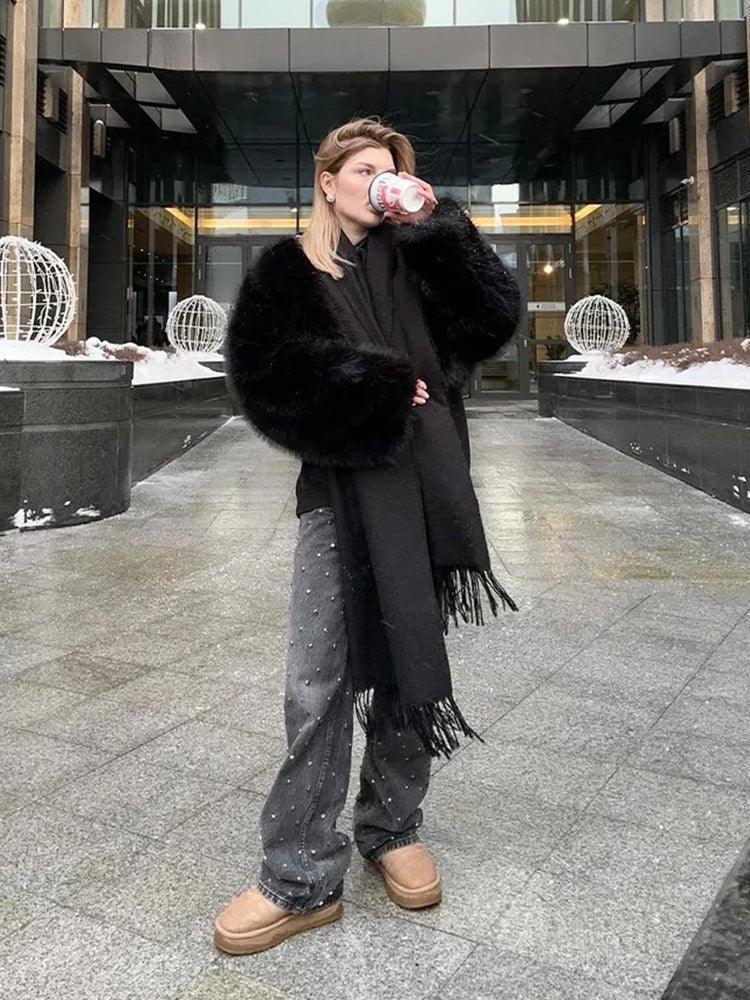 Woman wearing a black faux fur bomber jacket and scarf, drinking from a cup outdoors.