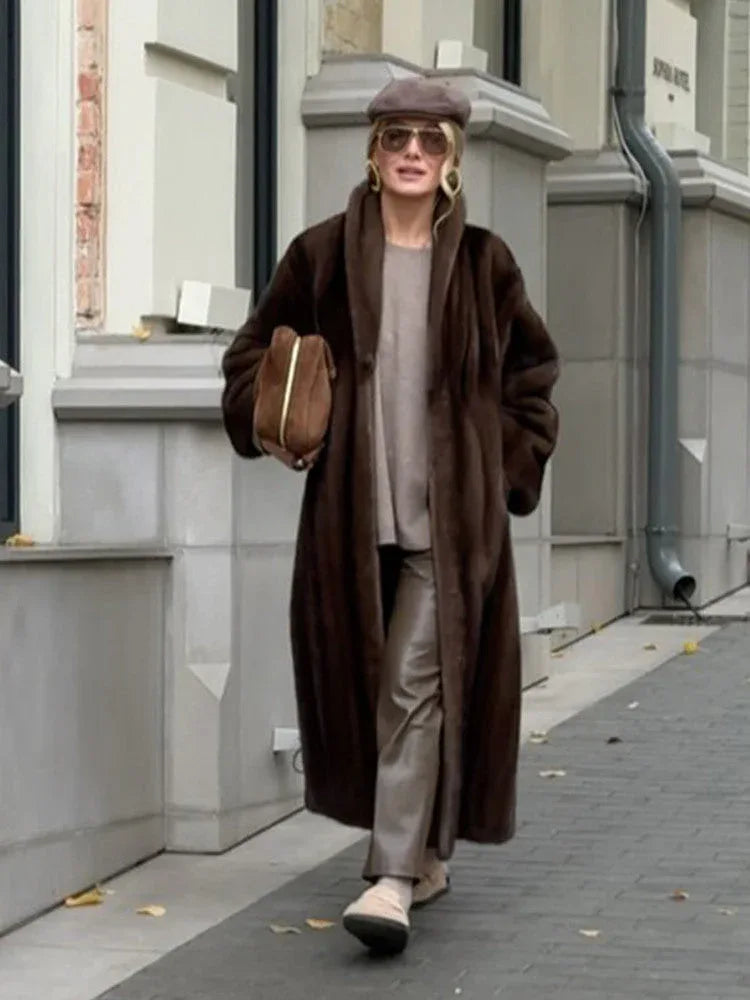 Woman wearing a long brown faux fur coat, a taupe knit sweater, taupe trousers, a taupe beret, sunglasses, and tan slip-on shoes, holding a brown suede handbag.