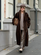 Woman wearing a long brown faux fur coat, a taupe knit sweater, taupe trousers, a taupe beret, sunglasses, and tan slip-on shoes, holding a brown suede handbag.