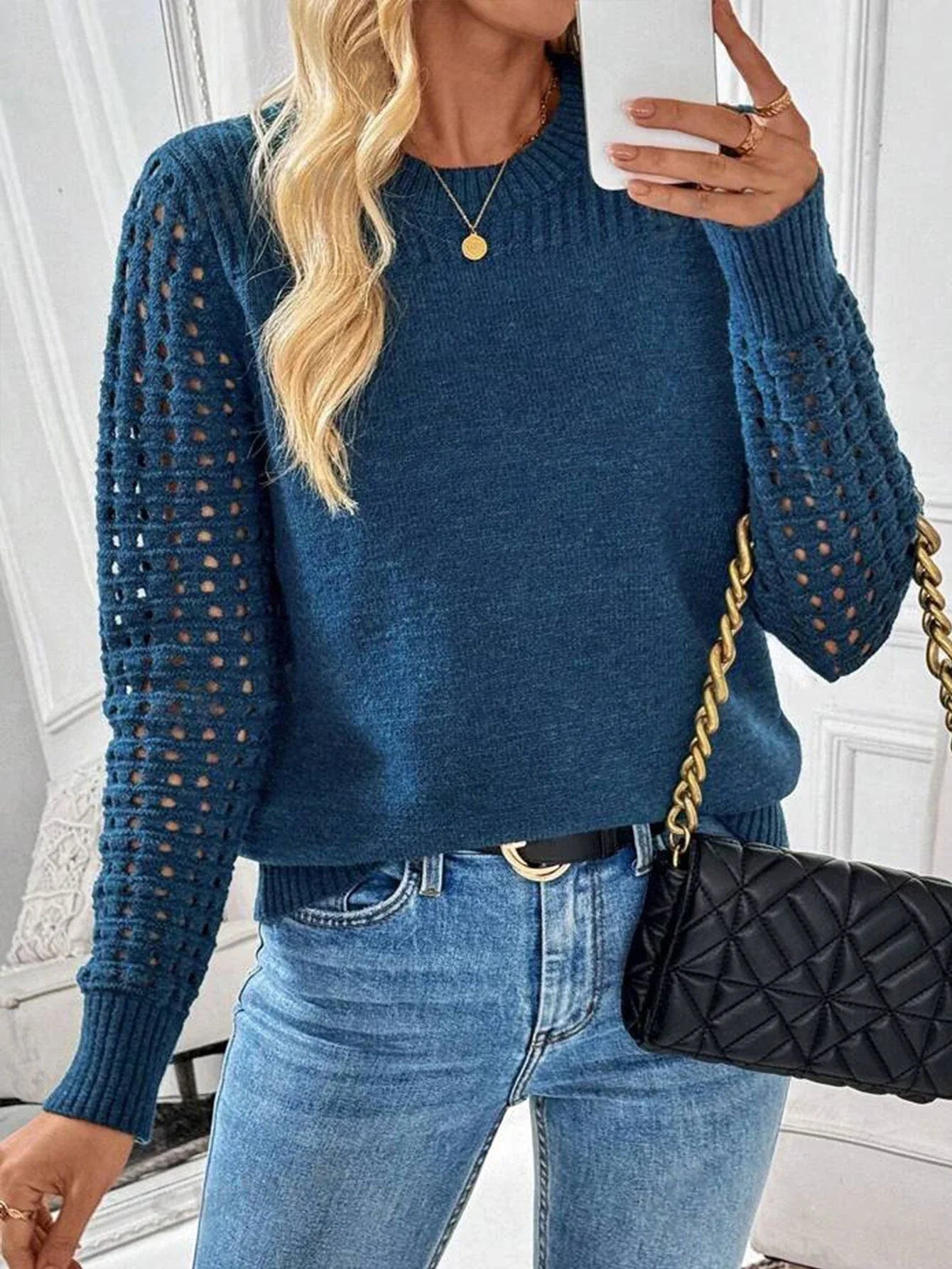 Woman holding a smartphone and handbag, wearing a blue knit sweater with openwork sleeves