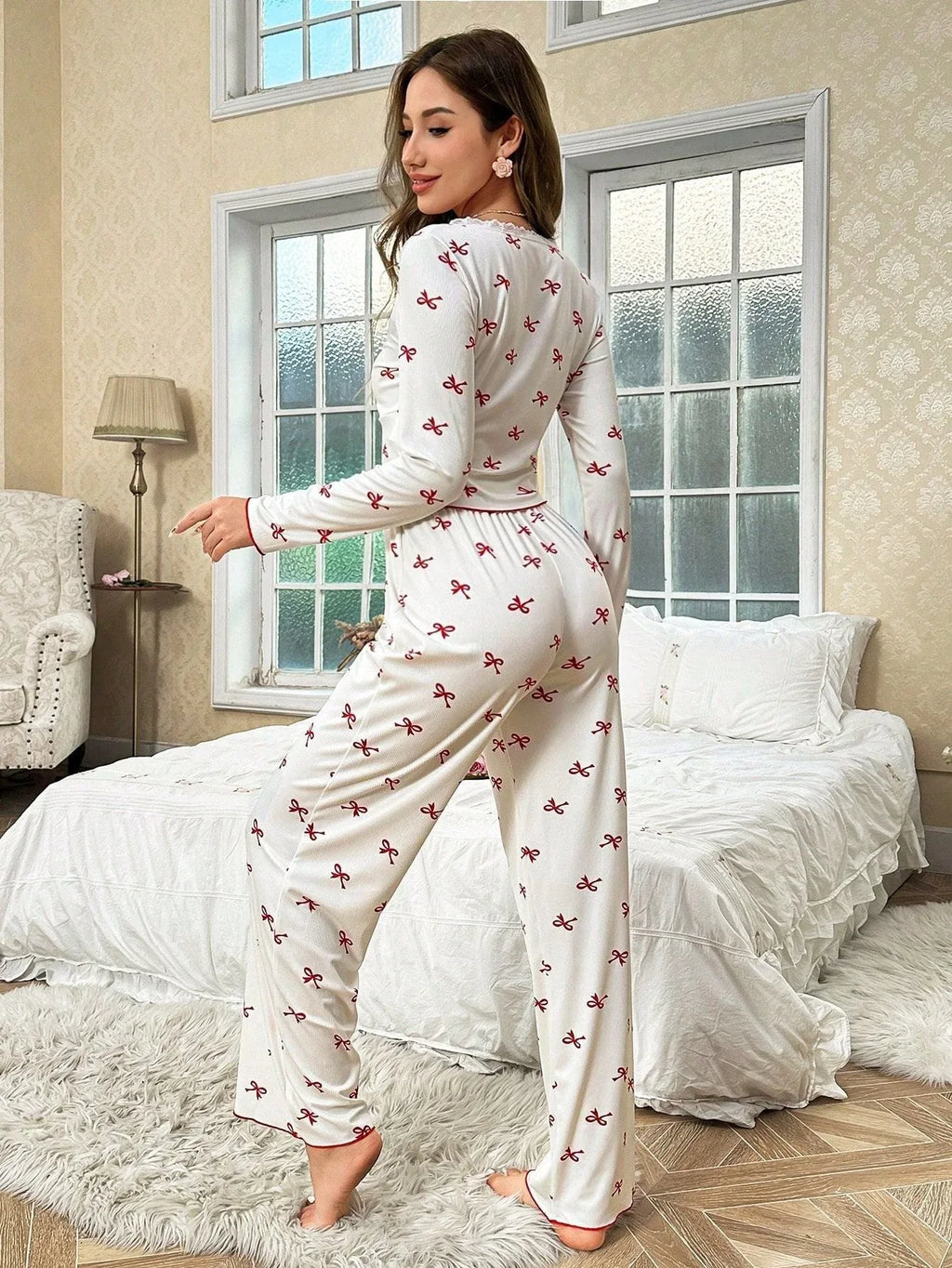 Woman wearing white long-sleeve pajama top with red bow print and matching pajama pants, posing in a bedroom.