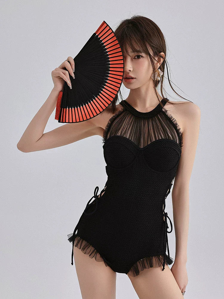 Woman wearing a black one-piece swimsuit with a sheer mesh halter neckline and ruffled hem, holding a red and black fan to her face.