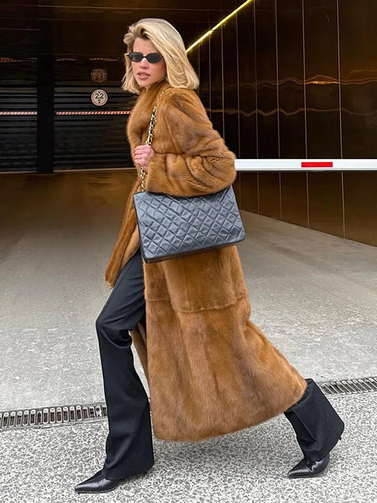Woman in a long brown faux fur coat walking with a black quilted handbag.