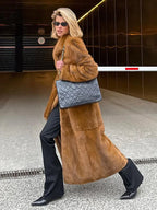 Woman in a long brown faux fur coat walking with a black quilted handbag.