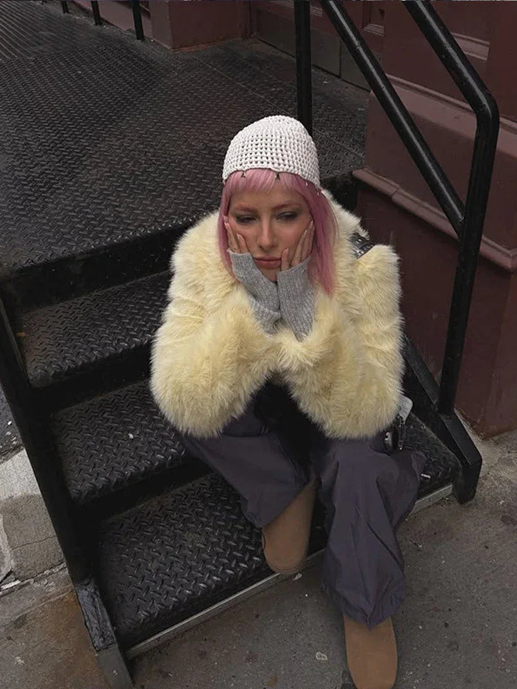 Woman sitting on stairs wearing a cream faux fur cropped jacket with a lapel collar and long sleeves, paired with grey pants and a grey turtleneck.