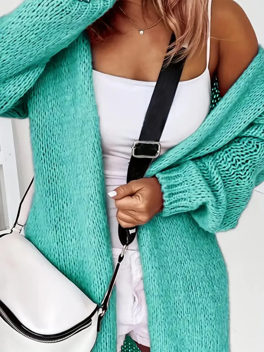 Close-up of a woman wearing a longline teal chunky knit cardigan with a white tank top and white shorts, carrying a white crossbody bag.