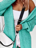 Close-up of a woman wearing a longline teal chunky knit cardigan with a white tank top and white shorts, carrying a white crossbody bag.