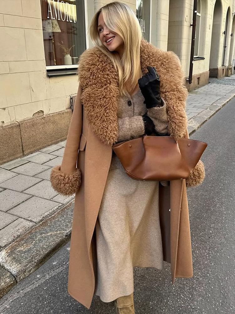Woman wearing a long camel wool coat with a fluffy shearling collar and cuffs, holding a brown handbag. Main image