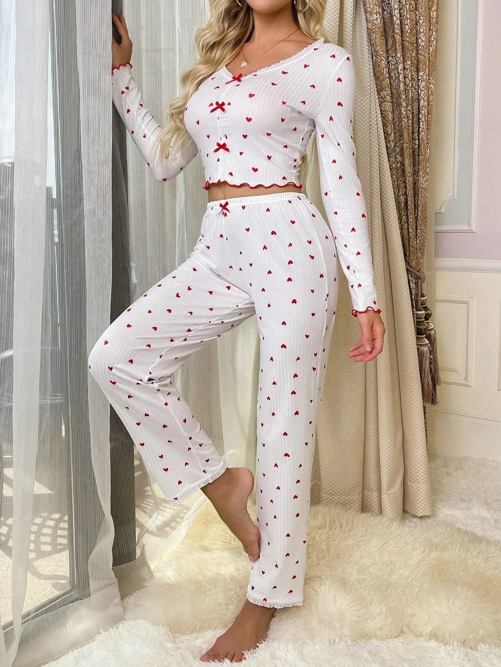 Woman leaning against a window frame, wearing a white pajama set with a red heart print and red bows.