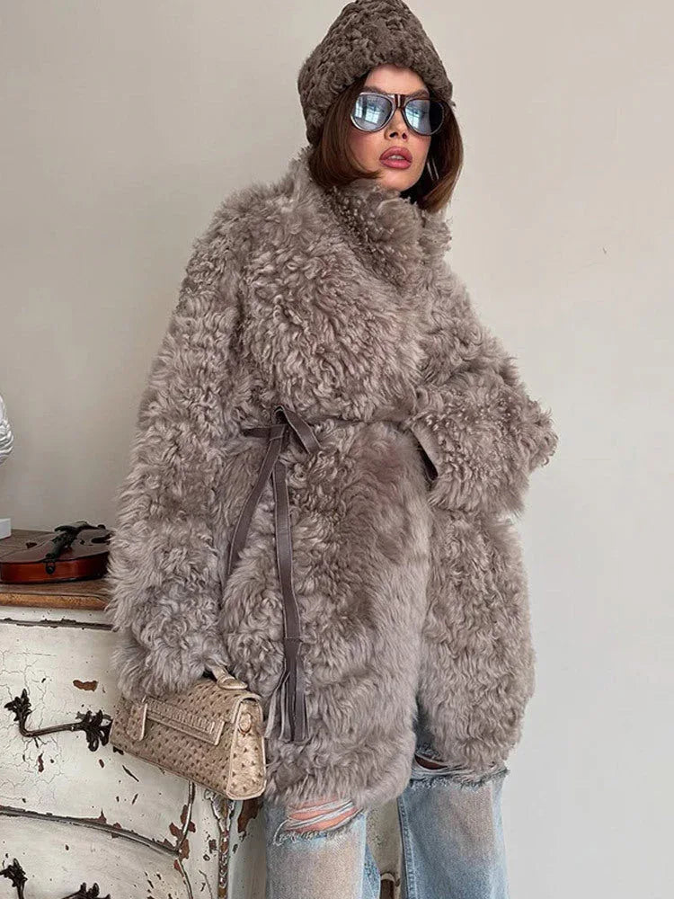 Woman demonstrating the belted feature of a grey curly faux fur coat, tied at the waist with a dark leather belt.