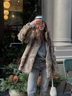 Woman wearing a brown and tan faux fur jacket with grey jeans and a green baseball cap, holding a coffee cup.
