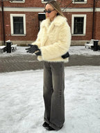 Woman wearing a cream faux fur jacket with wide lapels, grey wide-leg jeans, and black gloves, standing in the snow.