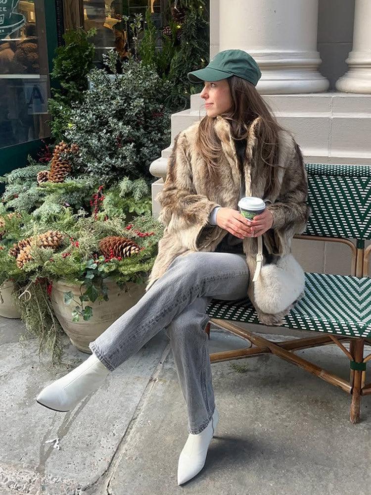 Woman sitting on a bench wearing a brown and tan faux fur jacket, grey jeans, and white ankle boots, holding a coffee cup.