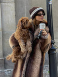 Woman holding a small dog while wearing a long, striped faux fur coat and a knit hat.