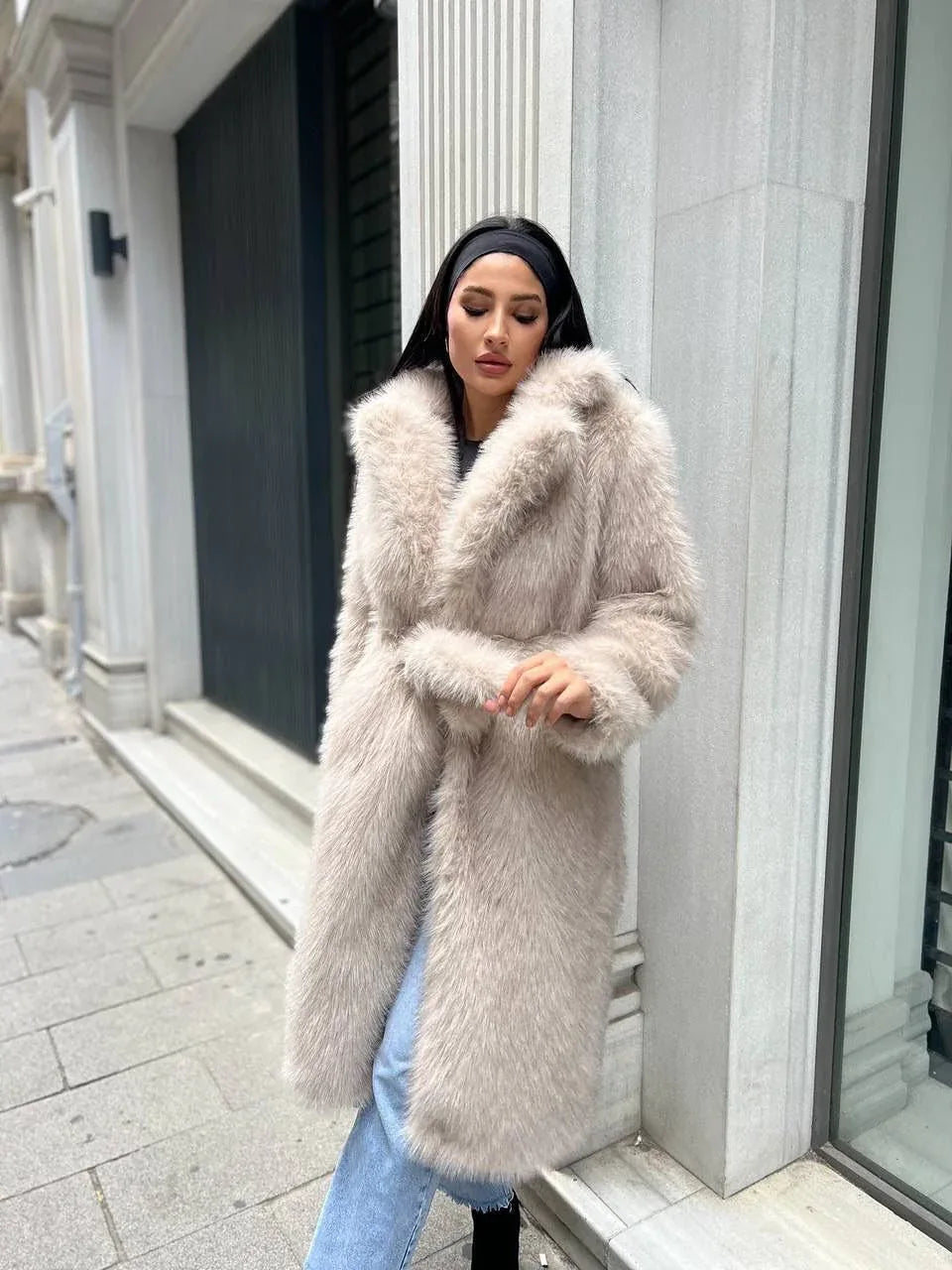 Woman wearing a long, light beige faux fur coat with a belted waist and lapel collar.