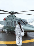 Woman in a long white and black spotted faux fur coat standing by a helicopter.
