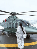 Woman in a long white and black spotted faux fur coat standing by a helicopter.