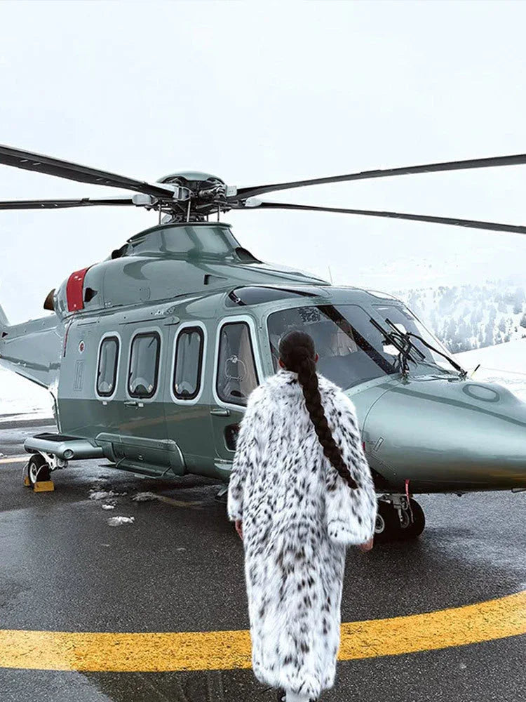 Woman in a long white and black spotted faux fur coat standing by a helicopter.