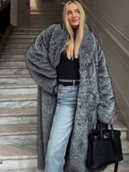 Woman wearing a long grey faux fur coat with a lapel collar and oversized sleeves, paired with blue jeans.