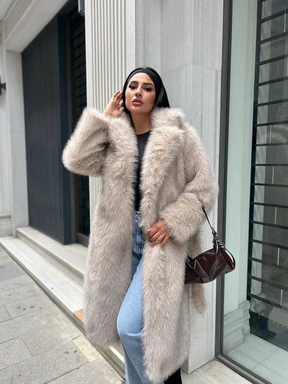 Woman in a light beige faux fur coat with a belted waist and lapel collar, holding a handbag.