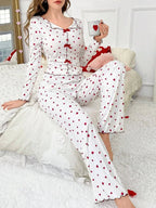 Woman sitting on a couch wearing a white long-sleeve pajama set with a red heart print and red trim, holding a red heart pillow.