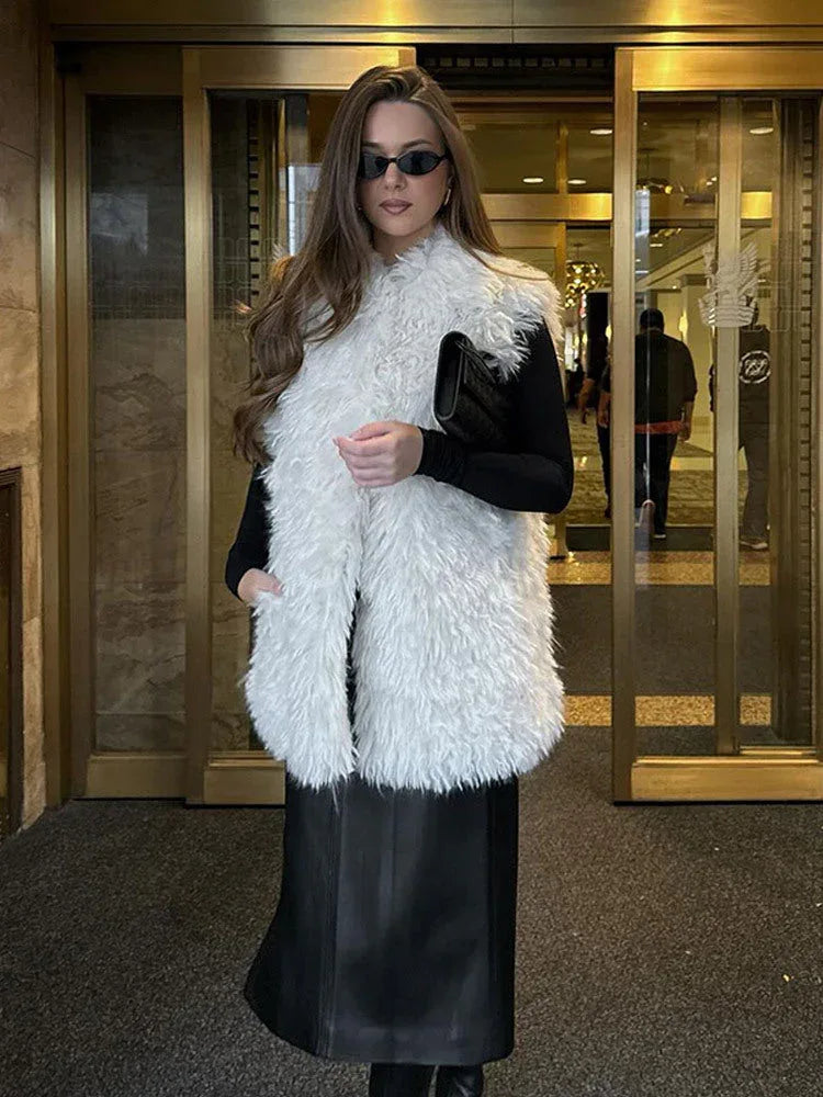 Woman wearing a white faux fur vest over a black long-sleeved top and a black midi skirt.