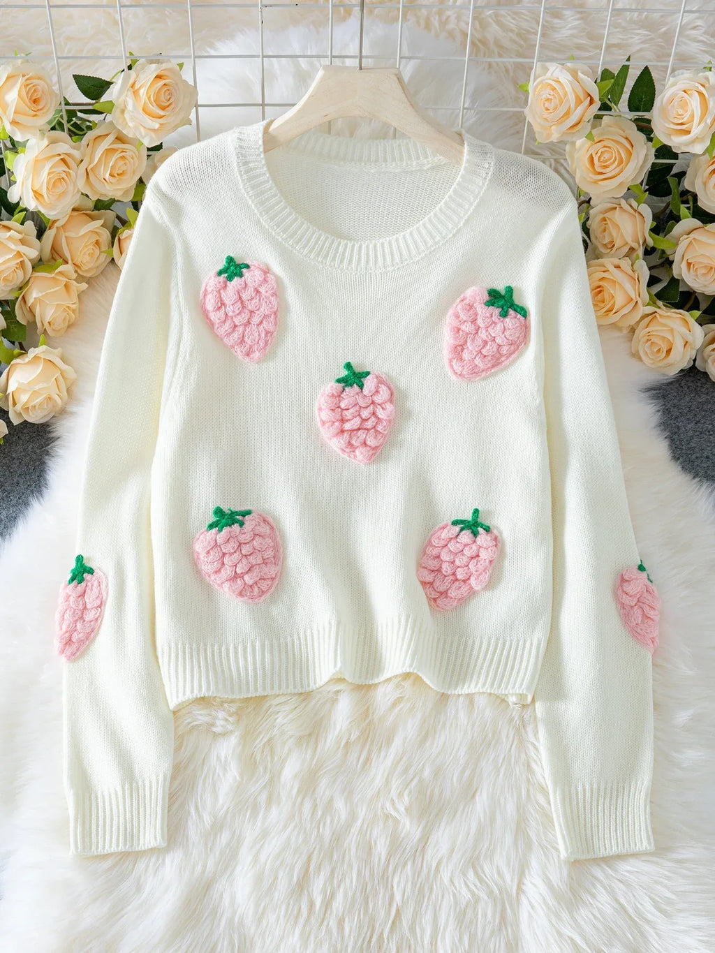 White cropped knit sweater with embroidered pink strawberry appliques and green stems, displayed on a hanger with a floral backdrop.