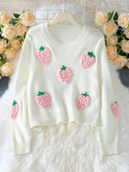 White cropped knit sweater with embroidered pink strawberry appliques and green stems, displayed on a hanger with a floral backdrop.