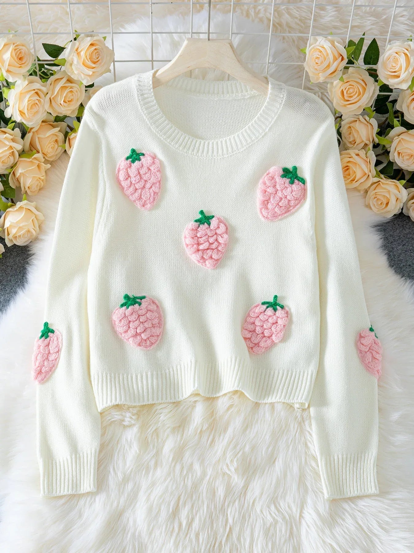 White cropped knit sweater with embroidered pink strawberry appliques and green stems, displayed on a hanger with a floral backdrop.