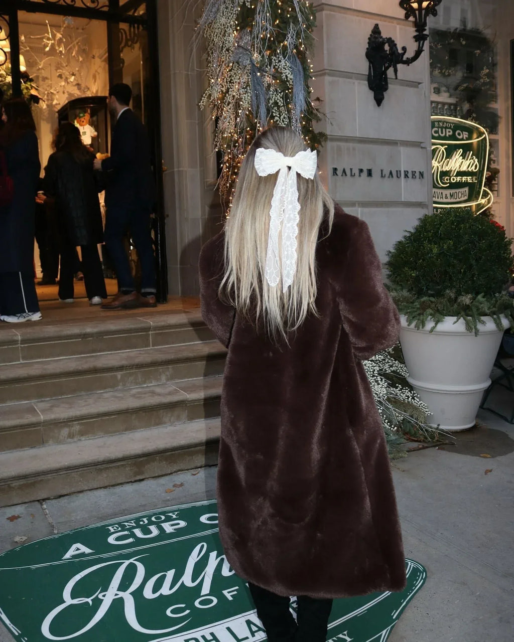 Woman from behind wearing a long brown faux fur coat with a large white bow in her hair