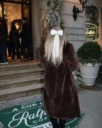 Woman from behind wearing a long brown faux fur coat with a large white bow in her hair