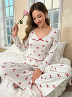 Woman holding a rose while wearing a white long-sleeve pajama top with red bow print and matching pajama pants, sitting on a bed.
