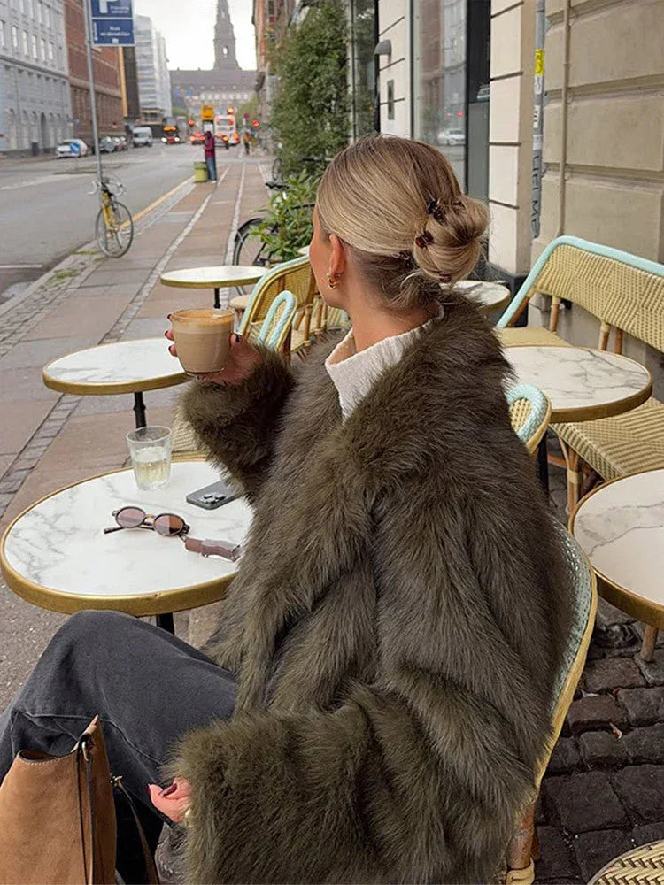 Woman from the side wearing a green faux fur jacket and white turtleneck, holding a coffee cup at an outdoor cafe.
