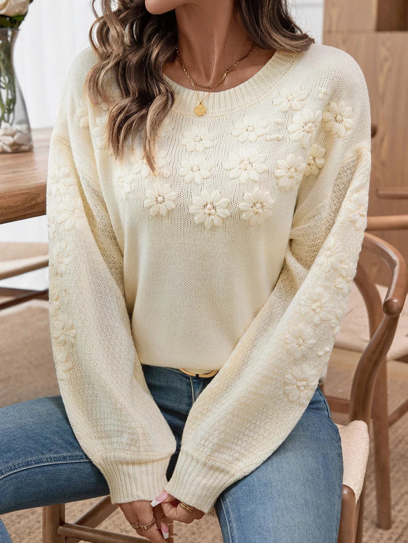 Woman seated, showcasing a cream knit sweater with floral embellishments and balloon sleeves.