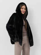 Woman wearing a black hooded faux fur jacket with a zipper closure and a taupe ribbed knit top.